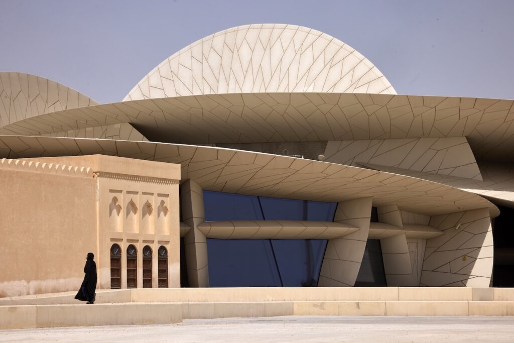 A person in a black abaya walks past the National Museum of Qatar, characterized by its distinctive, sand-colored, disk-shaped architecture reminiscent of a Rirkrit Tiravanija installation next to a traditional building.
