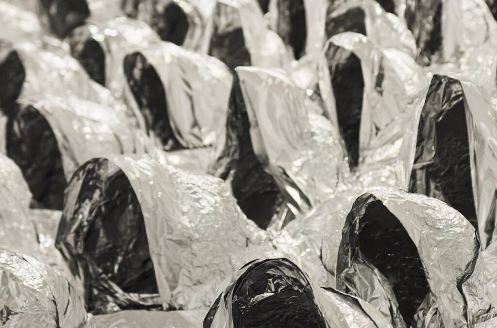 Dark objects by Rirkrit Tiravanija wrapped in wrinkled silver foil arranged in rows.