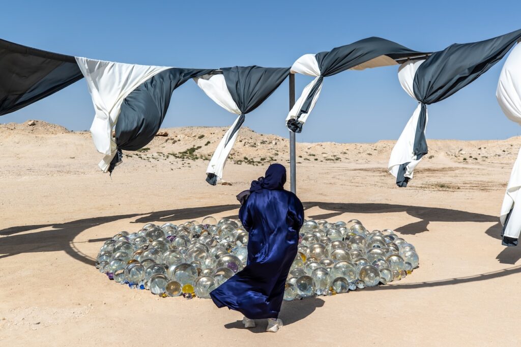A person in a blue abaya stands in a desert looking at Rirkrit Tiravanija’s circular cluster of glass spheres beneath draped black and white fabric.