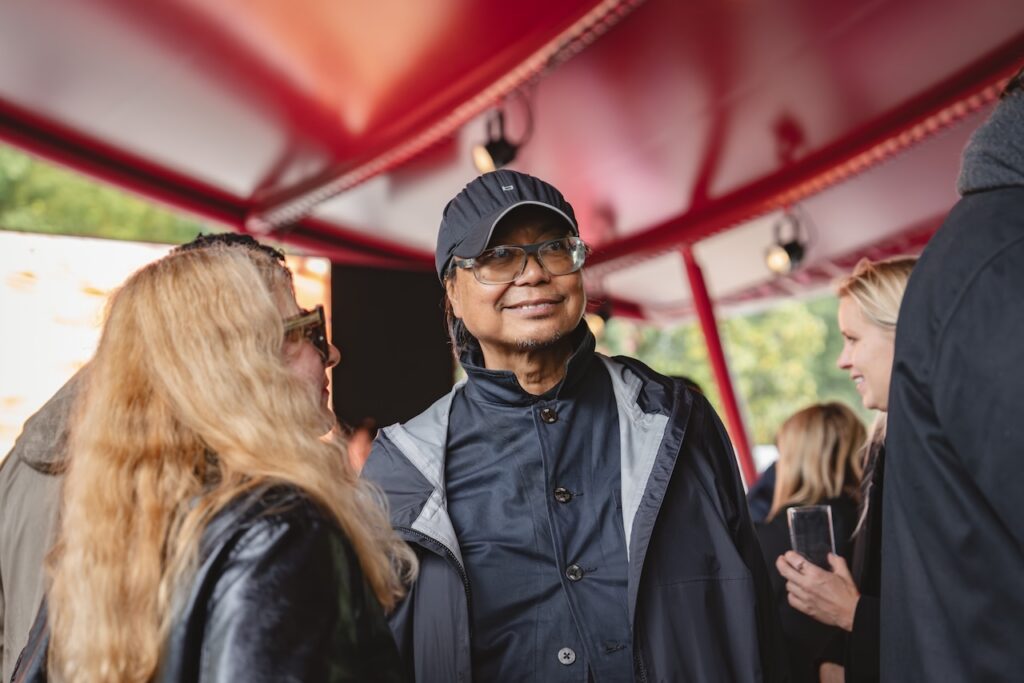 A person wearing a black cap and raincoat smiles while standing among a group of people under a red-roofed shelter