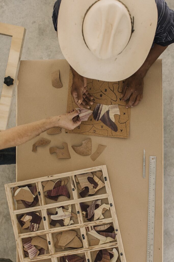 Two people, one of whom is wearing a hat, assemble a wooden puzzle at a table. A ruler and a compartment box with puzzle pieces sit nearby, all viewed from above.