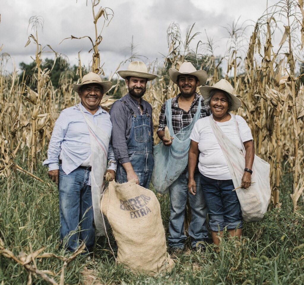 Four people in hats and casual clothes stand smiling in a cornfield, holding and carrying large sacks. corn plants are tall and dried, and the group appears happy and united.