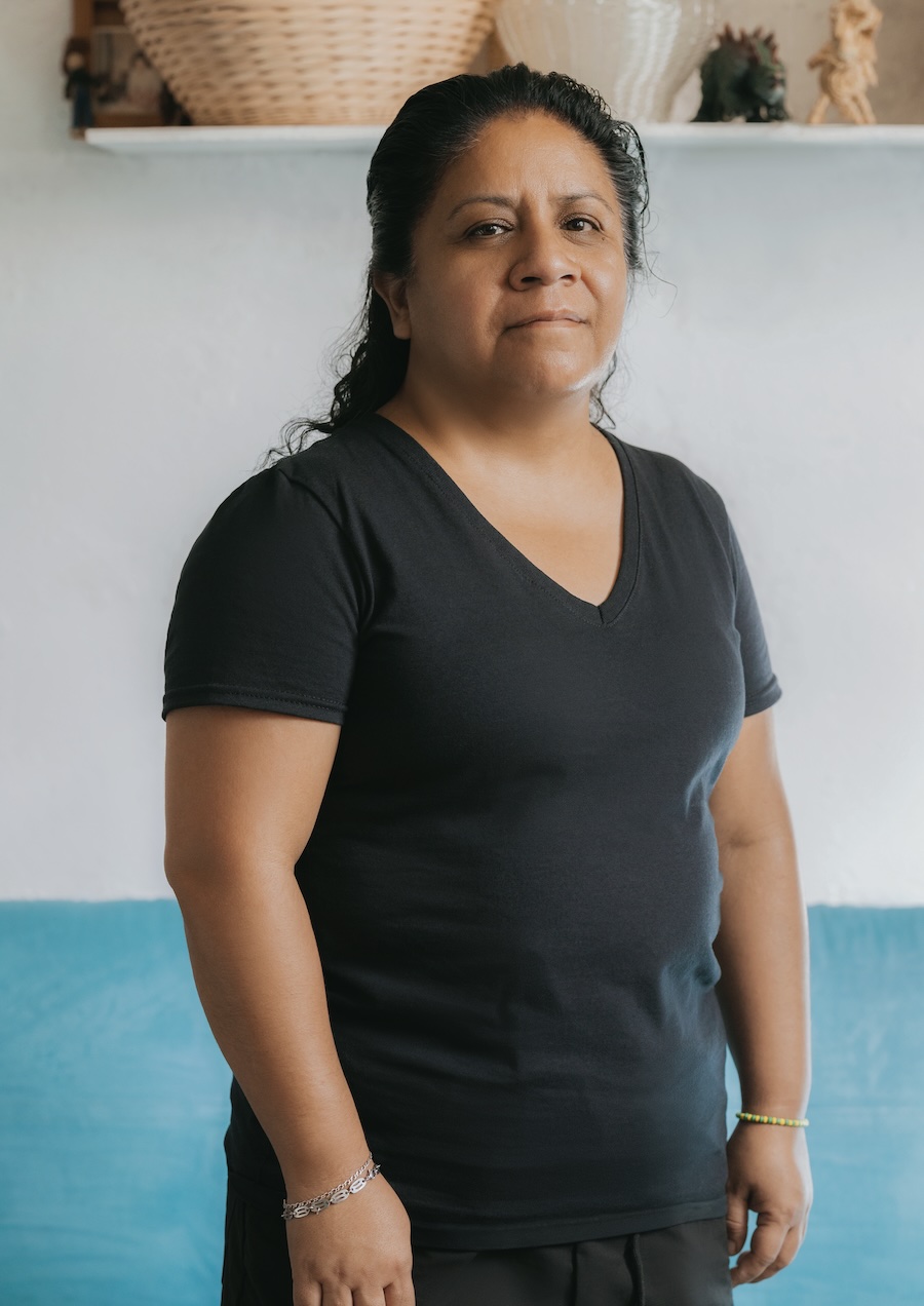 A woman with long dark hair wearing a black V-neck T-shirt stands indoors, looking at the camera with a neutral expression