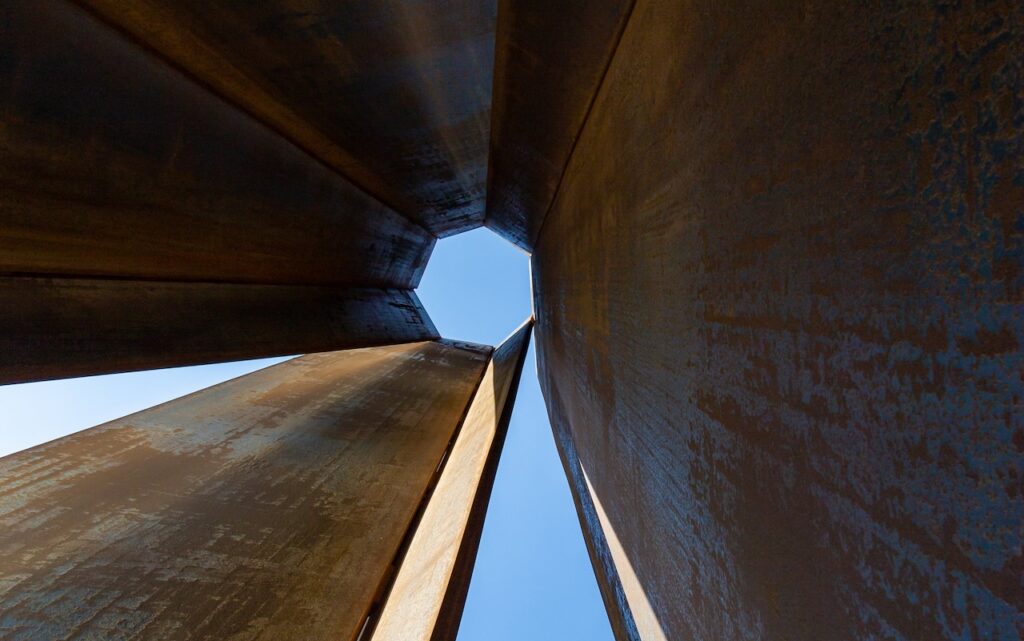 Looking up through the center of tall, rusted steel panels in the style of Rirkrit Tiravanija toward a patch of blue sky.