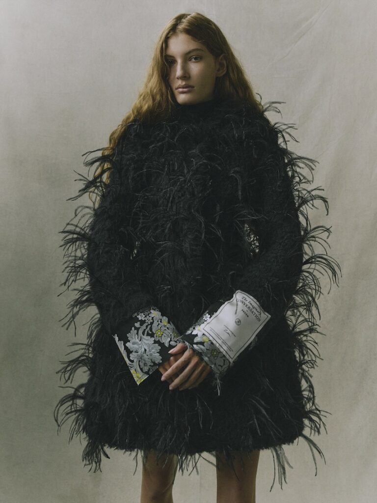 A person with long wavy hair wears a textured, feathered black coat with patterned cuffs, standing against a plain, light-colored background