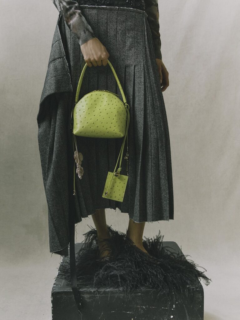 Person wearing a pleated gray skirt and feathered shoes stands on a black box, holding a textured lime green handbag and a small matching accessory, with a neutral backdrop behind them