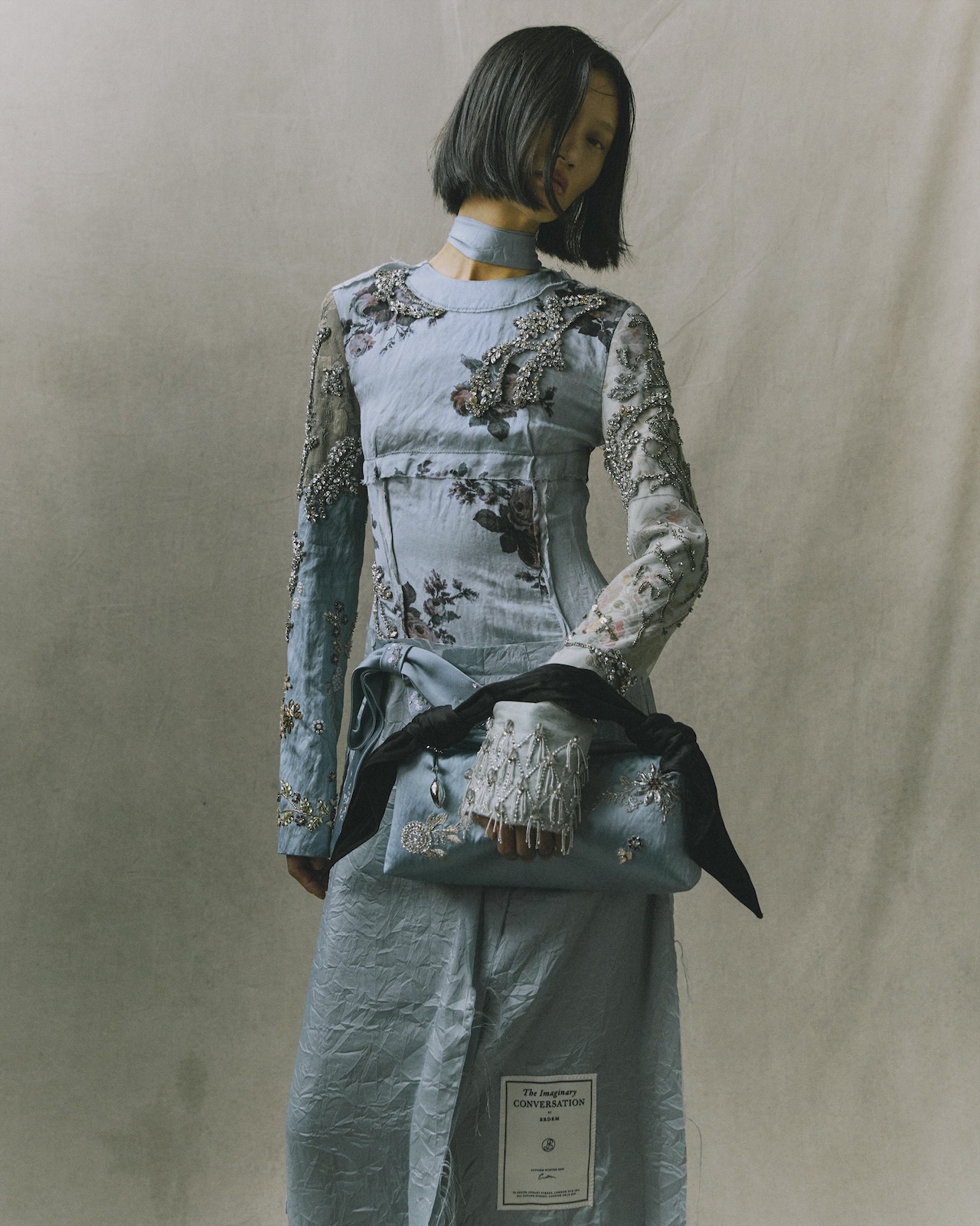 A person wearing an Erdem Moralioglu blue floral-print dress with beaded sleeve detailing and a matching blue clutch bag.