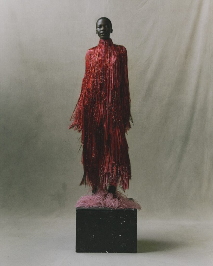 A person stands on a black pedestal wearing a long, red fringed dress with matching pink fringed shoes, against a neutral background.