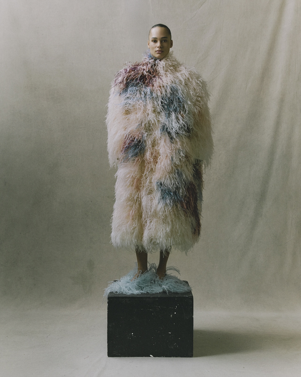 A model wearing a voluminous Erdem Moralioglu coat covered in pink, blue, and white feathers stands on a black block.