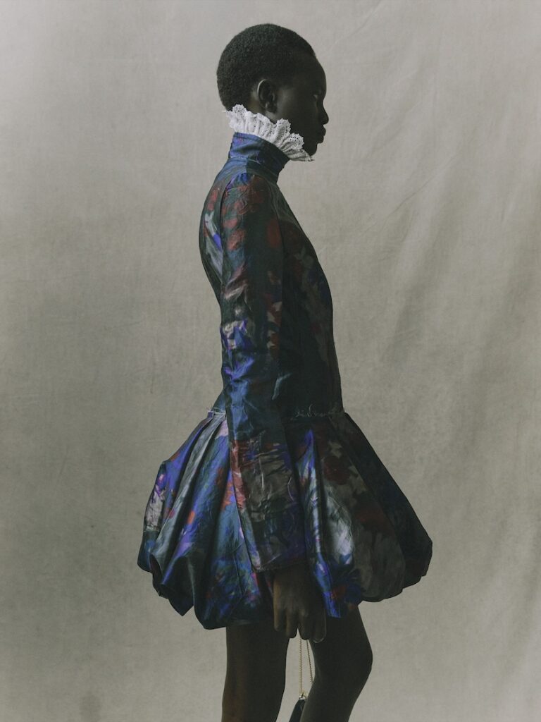 A person stands in profile wearing a blue, floral-patterned dress with a voluminous skirt and a white ruffled collar, against a plain, light background