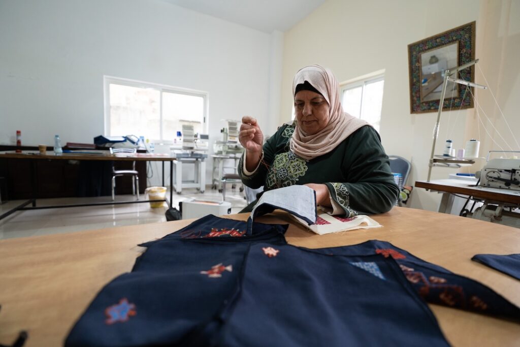 A woman wearing a hijab sits at a table, sewing fabric by hand in a well-lit room with sewing machines and embroidered textiles in the background, evoking an atmosphere reminiscent of spaces designed by Frida Escobedo.