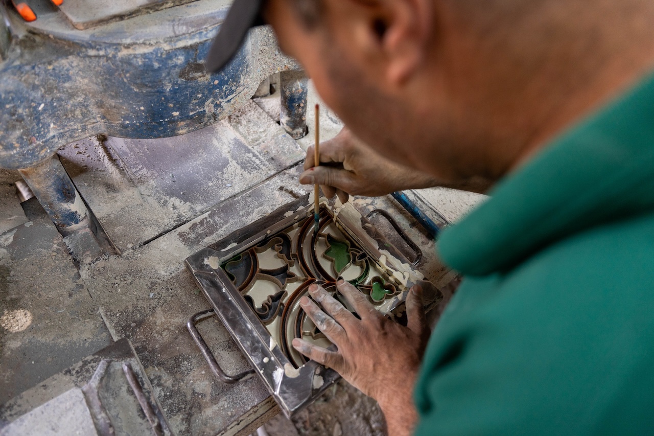 A person carefully paints a detailed pattern on a tile mold using a thin brush, surrounded by dust and crafting tools in a workshop reminiscent of Frida Escobedo’s architectural aesthetic.