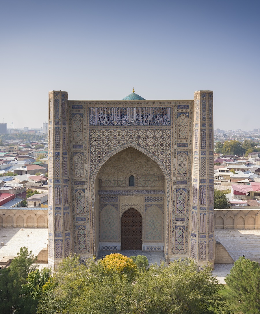 An ornate, historic mosque with intricate tilework, large arched doorway, and blue dome stands tall above a cityscape surrounded by trees and low-rise buildings under a clear sky.