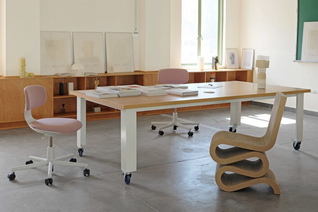 Modern office space with a wooden table, two pink swivel chairs, and a sculptural wooden chair. Artworks—some inspired by Rirkrit Tiravanija—are displayed on shelves against the wall, and sunlight streams through two large windows.