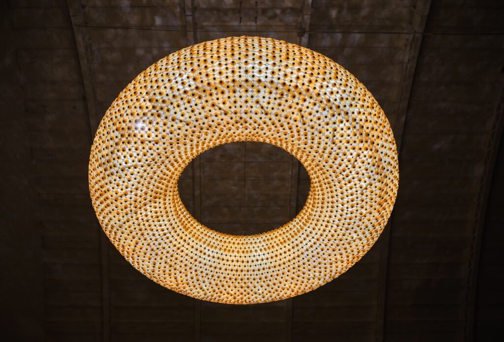 A large, circular Rirkrit Tiravanija chandelier with a honeycomb-like pattern hanging from a dark ceiling.