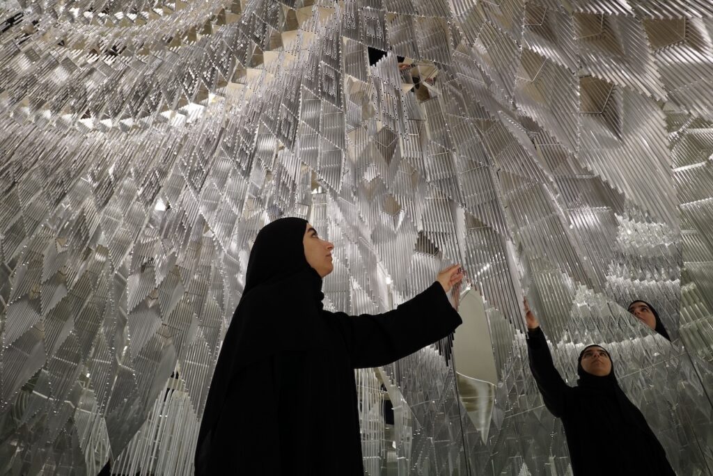 A woman in a black abaya touches a mirrored, geometric art installation, her reflection visible on the shiny surfaces surrounding her.