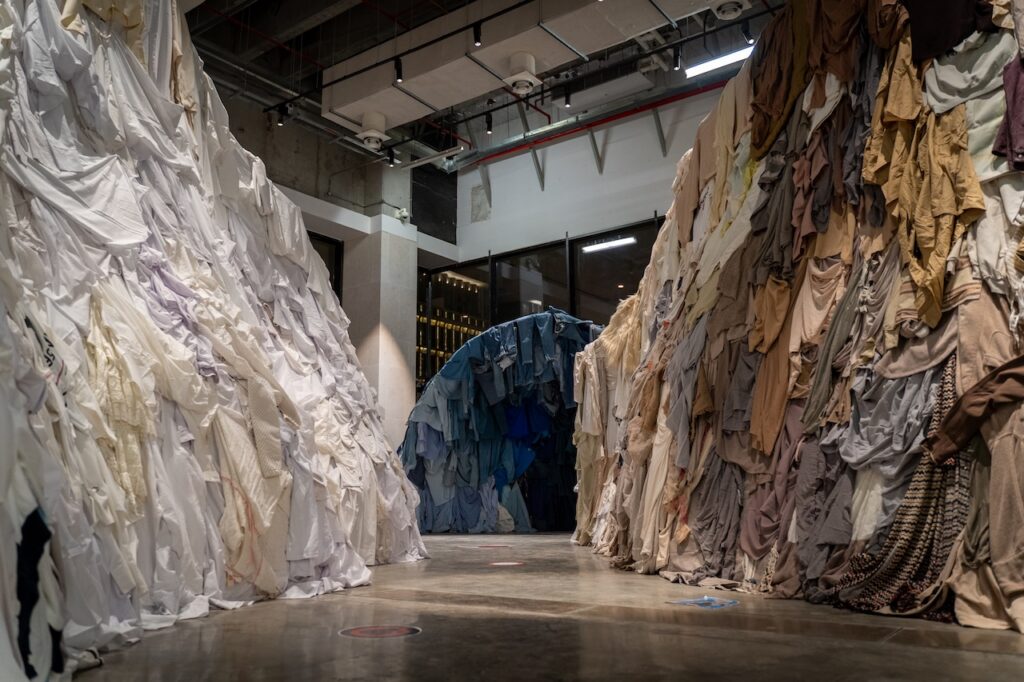 An art installation by Rirkrit Tiravanija consisting of two towering walls of draped fabrics, one side white and one side varied earth tones, with a blue fabric tunnel at the end of the walkway.
