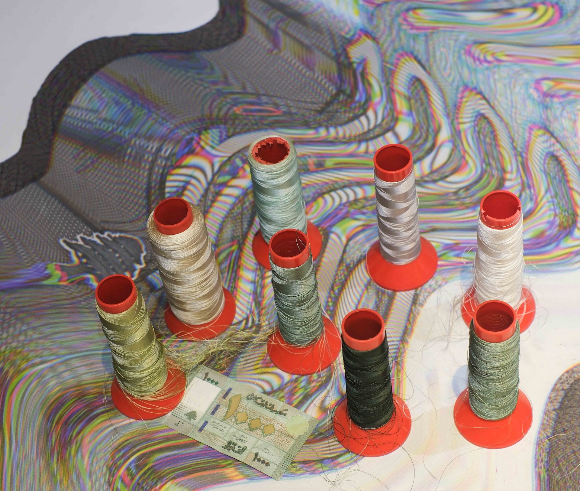 Eight spools of thread in various colors stand on red bases atop a wavy, multicolored surface with an Iranian banknote nearby.