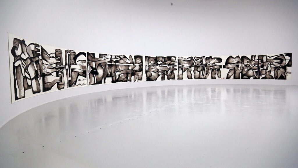A long, continuous black and white abstract ink painting by Rirkrit Tiravanija featuring organic, fragmented shapes, displayed on a curved white wall in a gallery.