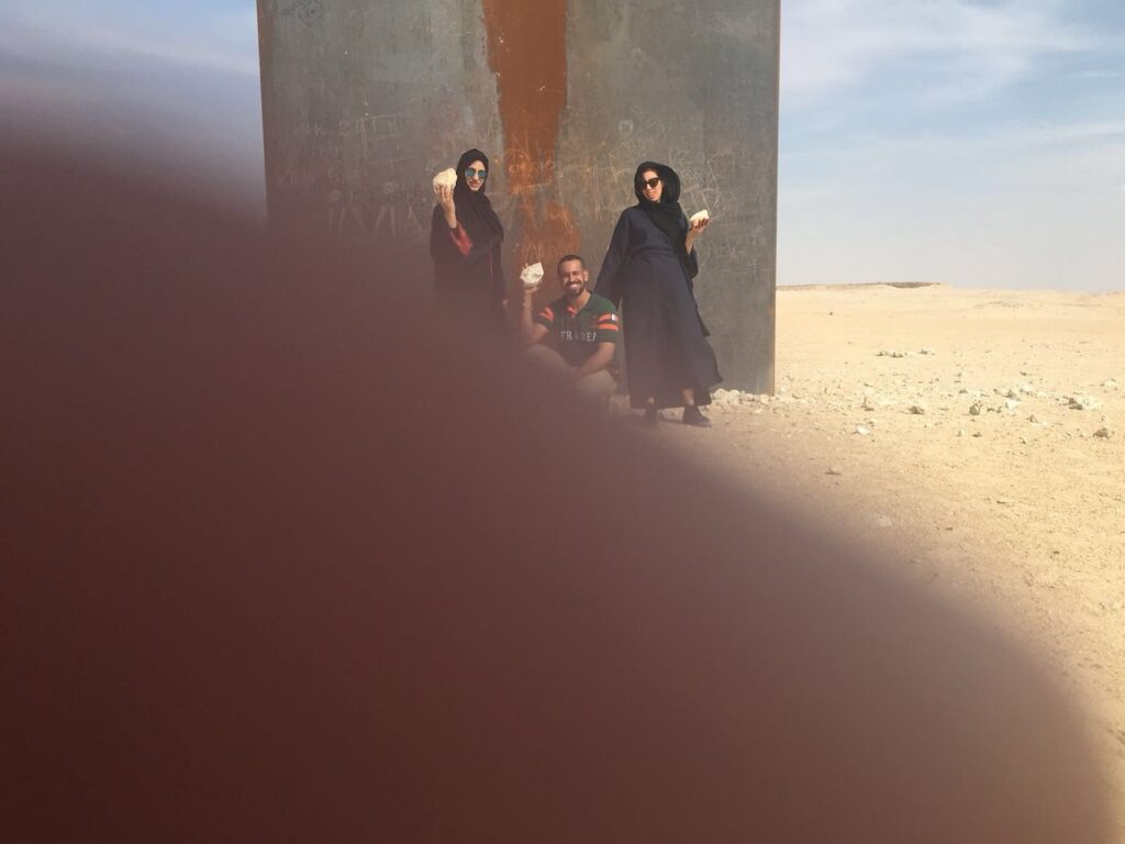 Three people pose in front of a rusty metal wall in a desert, holding objects. Two wear dark robes, one squats in the middle. The scene is partially obscured by a large thumb or finger covering the camera lens in the foreground.