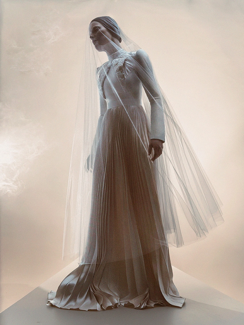 A person wearing a long, pleated, light-colored gown with long sleeves and a sheer veil stands against a soft, neutral background