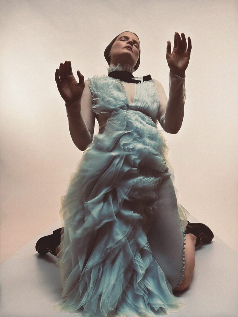 A person wearing a ruffled, light blue gown and sheer sleeves kneels with eyes closed and hands raised