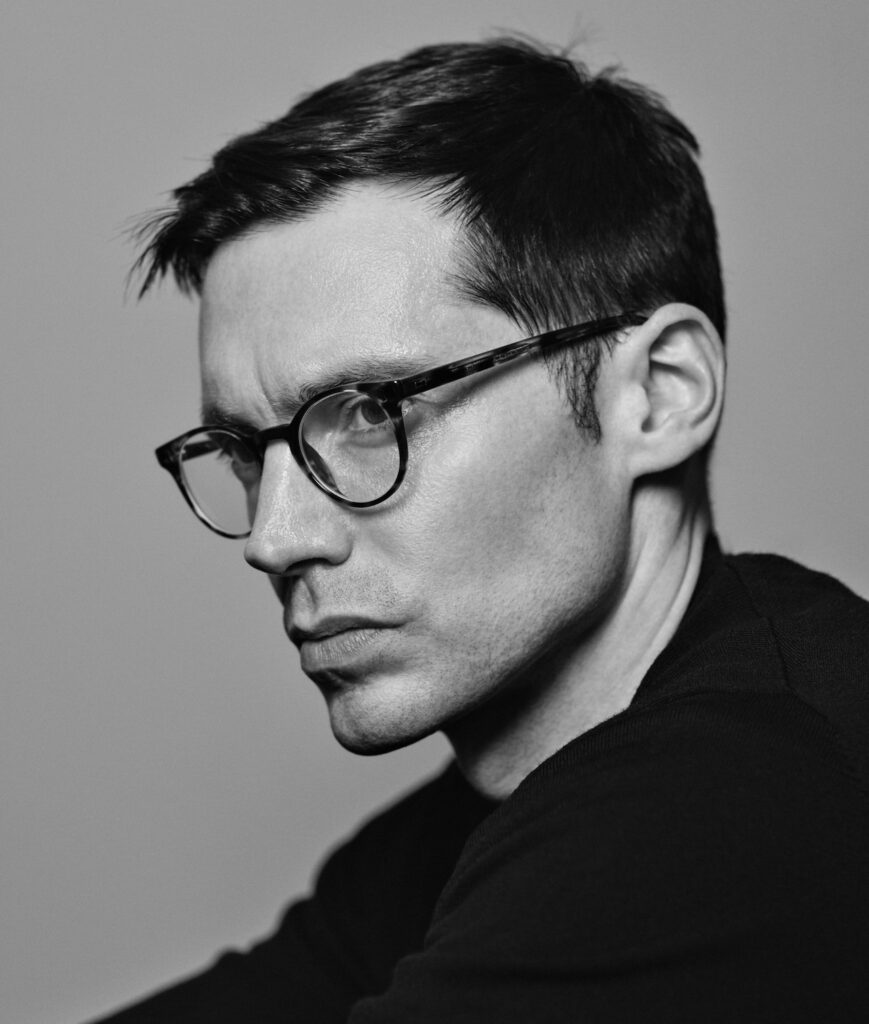 Black and white portrait of a man with short dark hair, wearing round glasses and a dark shirt, looking to the left with a serious expression against a plain background