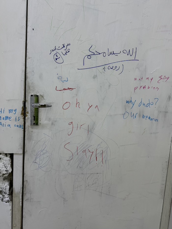 A white door covered in various handwritten messages and doodles in different colors, including “Oh ya girl slay!!” and “Hi my name is Ali,” along with writing in Arabic and other phrases