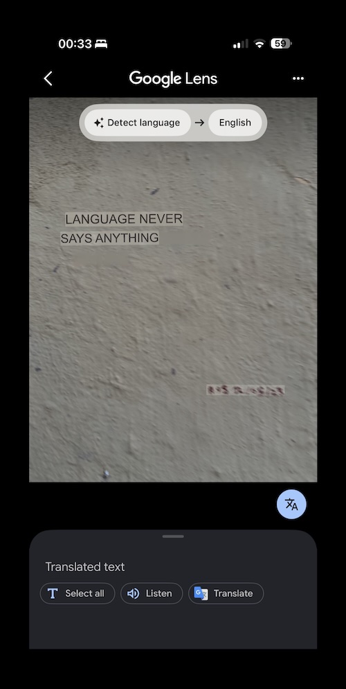 A phone screen displays Google Lens translating text on a rough beige wall that reads, LANGUAGE NEVER SAYS ANYTHING.