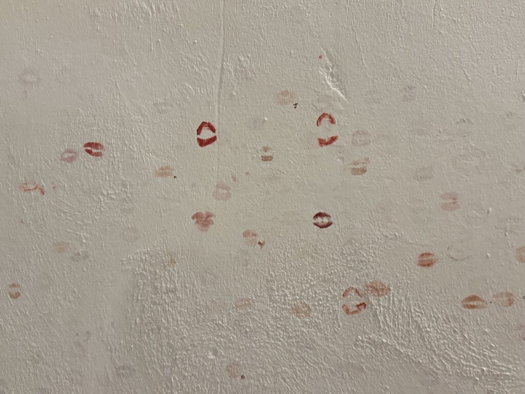 A white wall with multiple lipstick kiss marks in various shades of red and pink scattered across its textured surface