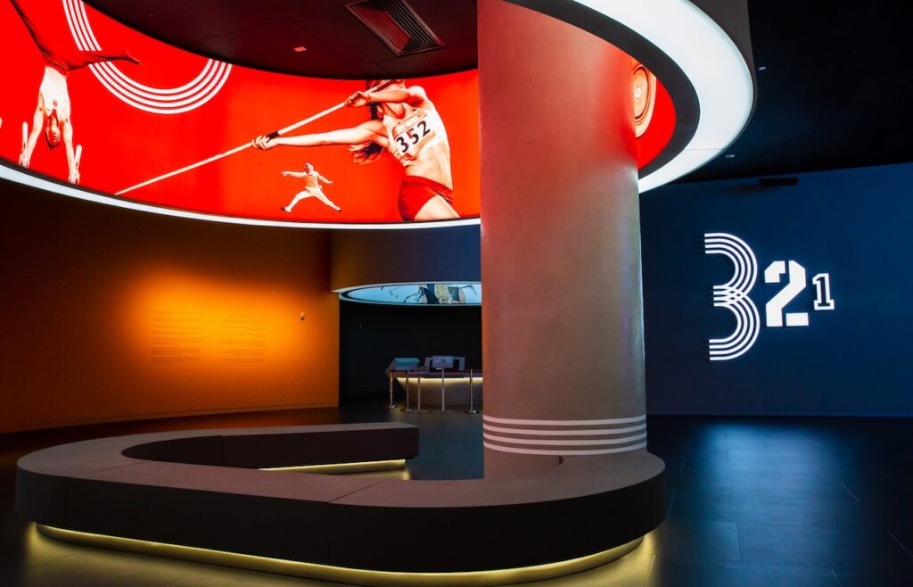 A curved Rirkrit Tiravanija interior features a circular red display screen showing athletes in motion, a central pillar with three white stripes, and a wall graphic displaying the numbers 3, 2, and 1.