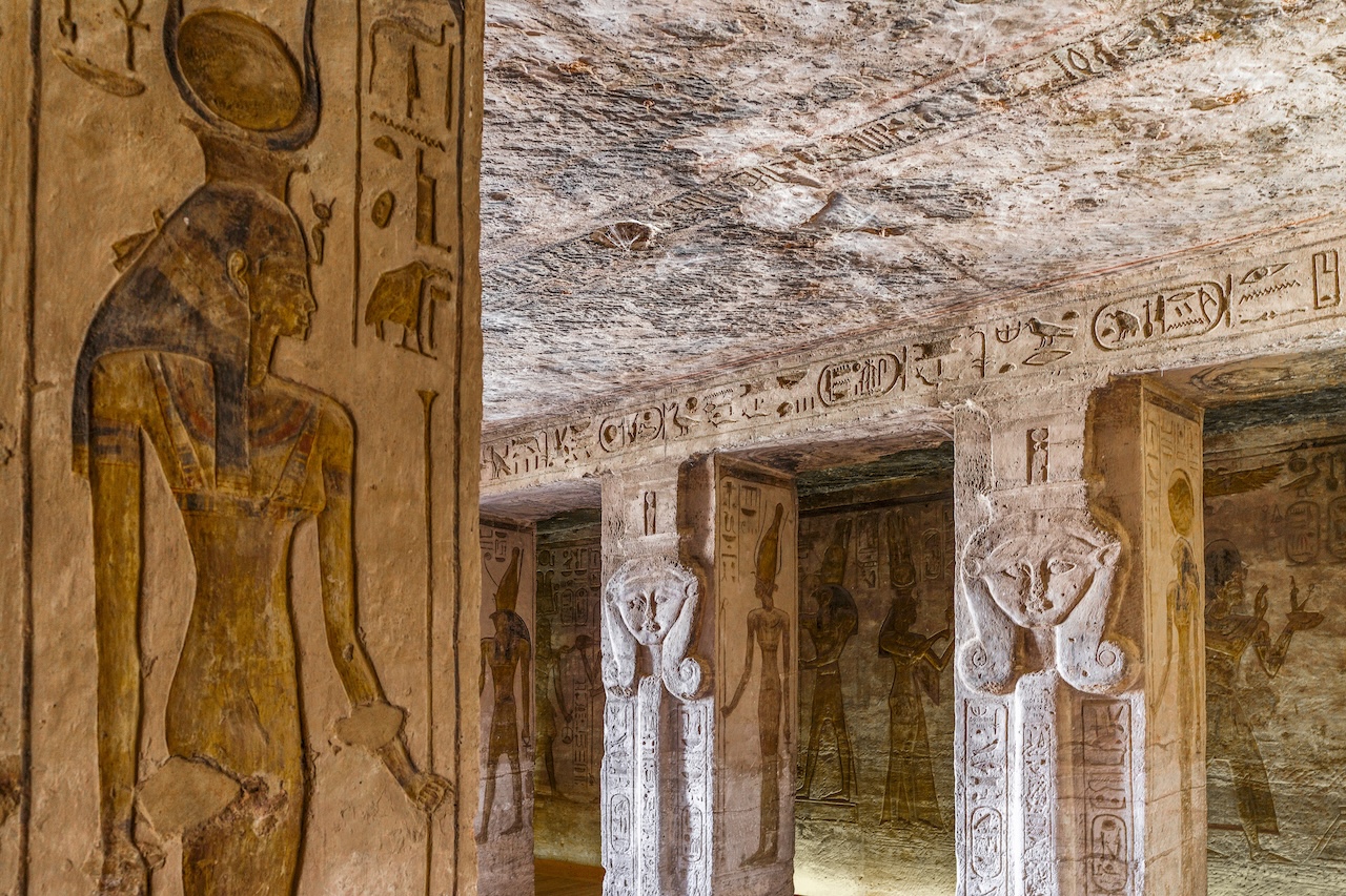 Intricate stone carvings of Egyptian deities and hieroglyphics, in the style documented by Luis Monreal, decorate the columns and walls inside an ancient temple.