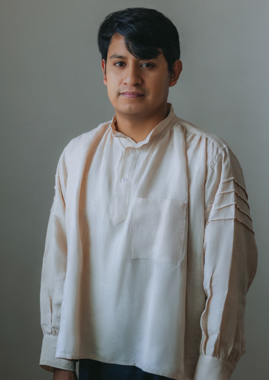 A young person with dark hair stands facing the camera, wearing a loose, light-colored shirt with decorative pleats on the sleeves