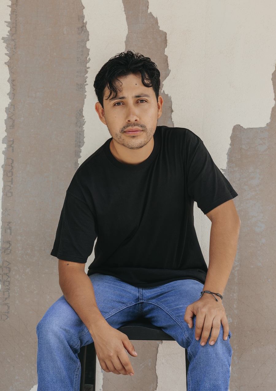A man with short dark hair and a beard, wearing a black t-shirt and blue jeans, sits on a stool in front of a textured, partially painted wall