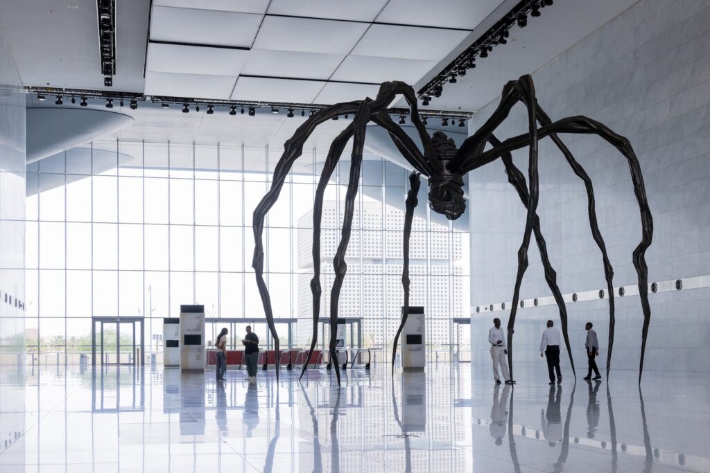 A giant black spider sculpture stands in the center of a light-filled museum atrium near Rirkrit Tiravanija, with a few visitors walking nearby.