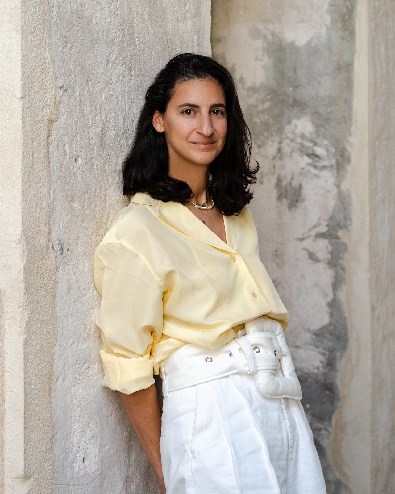 A woman with dark hair, wearing a light yellow blouse and high-waisted white pants, stands against a textured, light-colored wall, smiling softly at the camera
