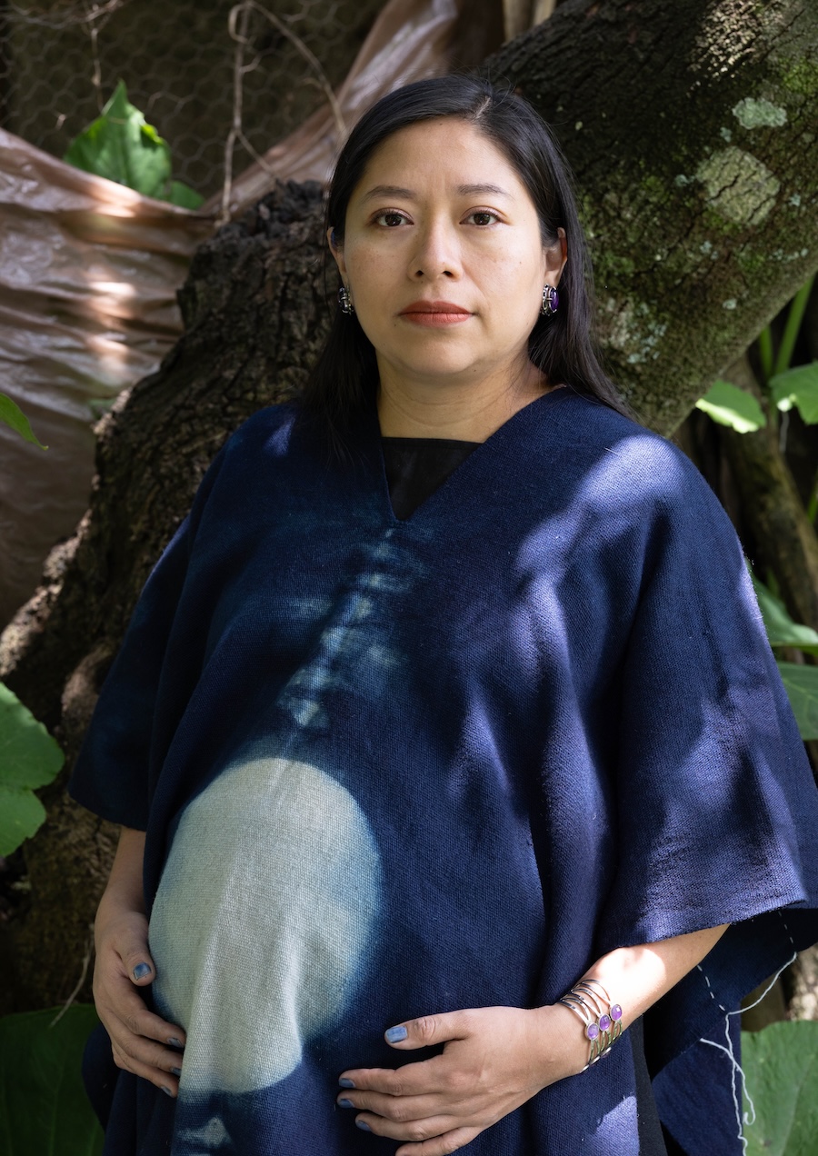 A woman with long dark hair, wearing a blue garment with a circular design, stands outdoors by a tree