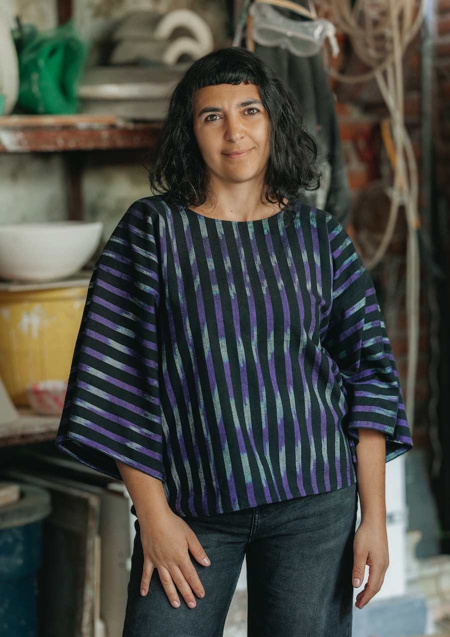 Awoman with dark hair and bangs, stands indoors wearing a black shirt with blue and green stripes and dark jeans. She poses casually, with shelves and pottery in the background.