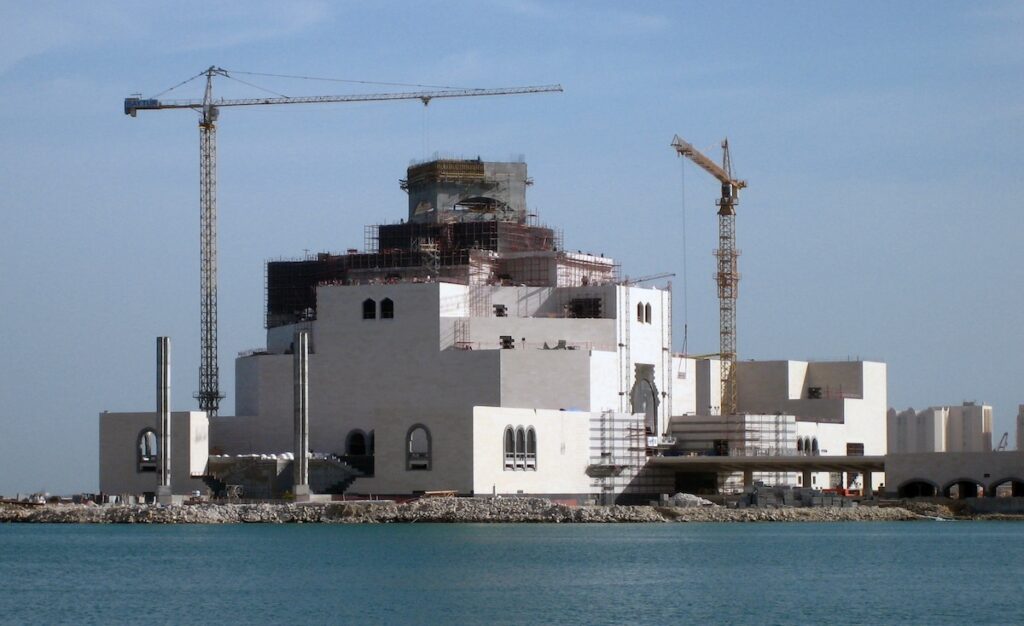 Large building under construction with two cranes on the waterfront, in the style of Rirkrit Tiravanija.