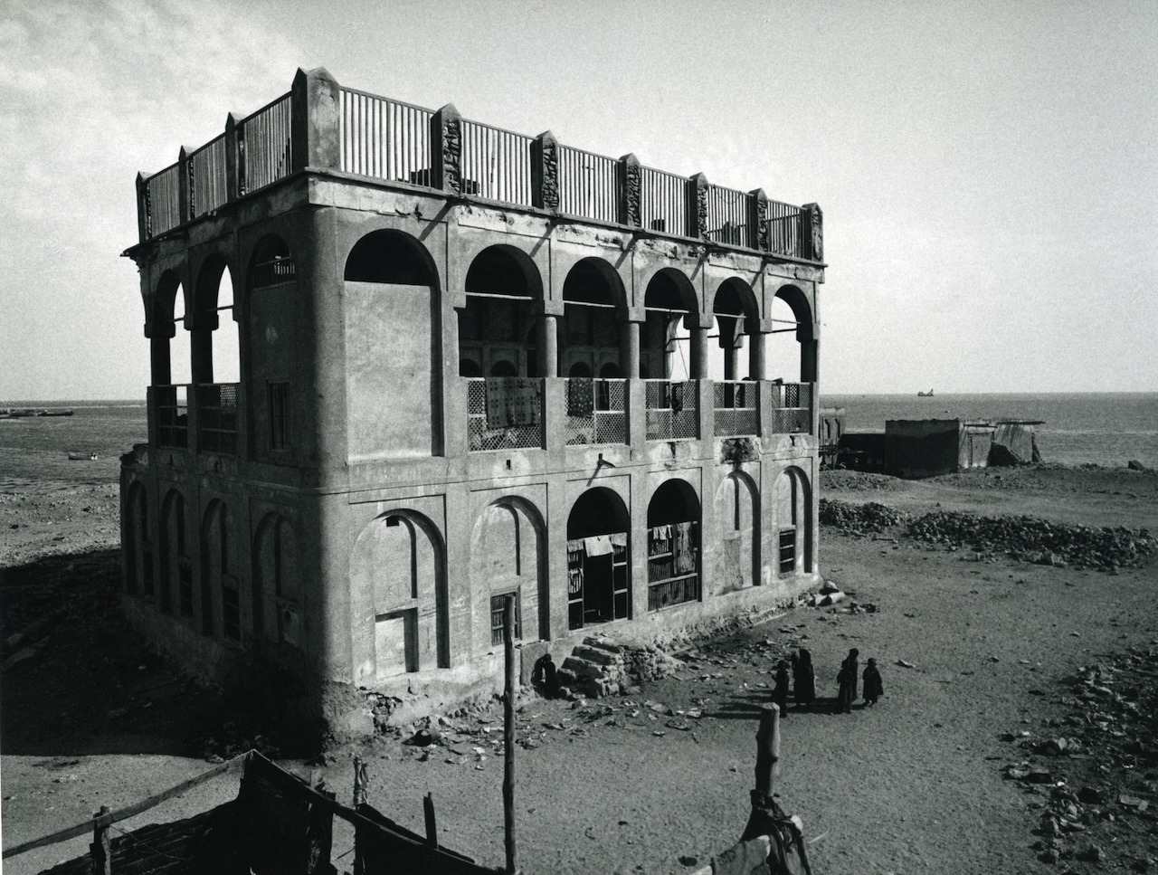 A large, two-story, abandoned building with arches stands in a barren, rocky landscape near the sea. A few people are gathered near the base.The photo is in black and white.