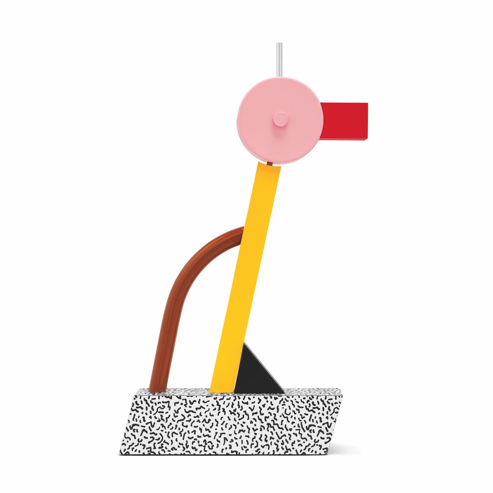 Abstract sculpture with geometric shapes: a speckled white base, upright yellow and brown rods, a pink circle with red and gray accents on top, and a small black triangle at the bottom.