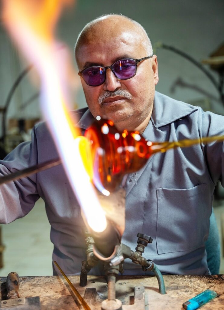 A man wearing tinted glasses shapes hot glass with a metal rod over a flame, working intently in a workshop reminiscent of the innovative spirit seen in Frida Escobedo’s architectural designs.