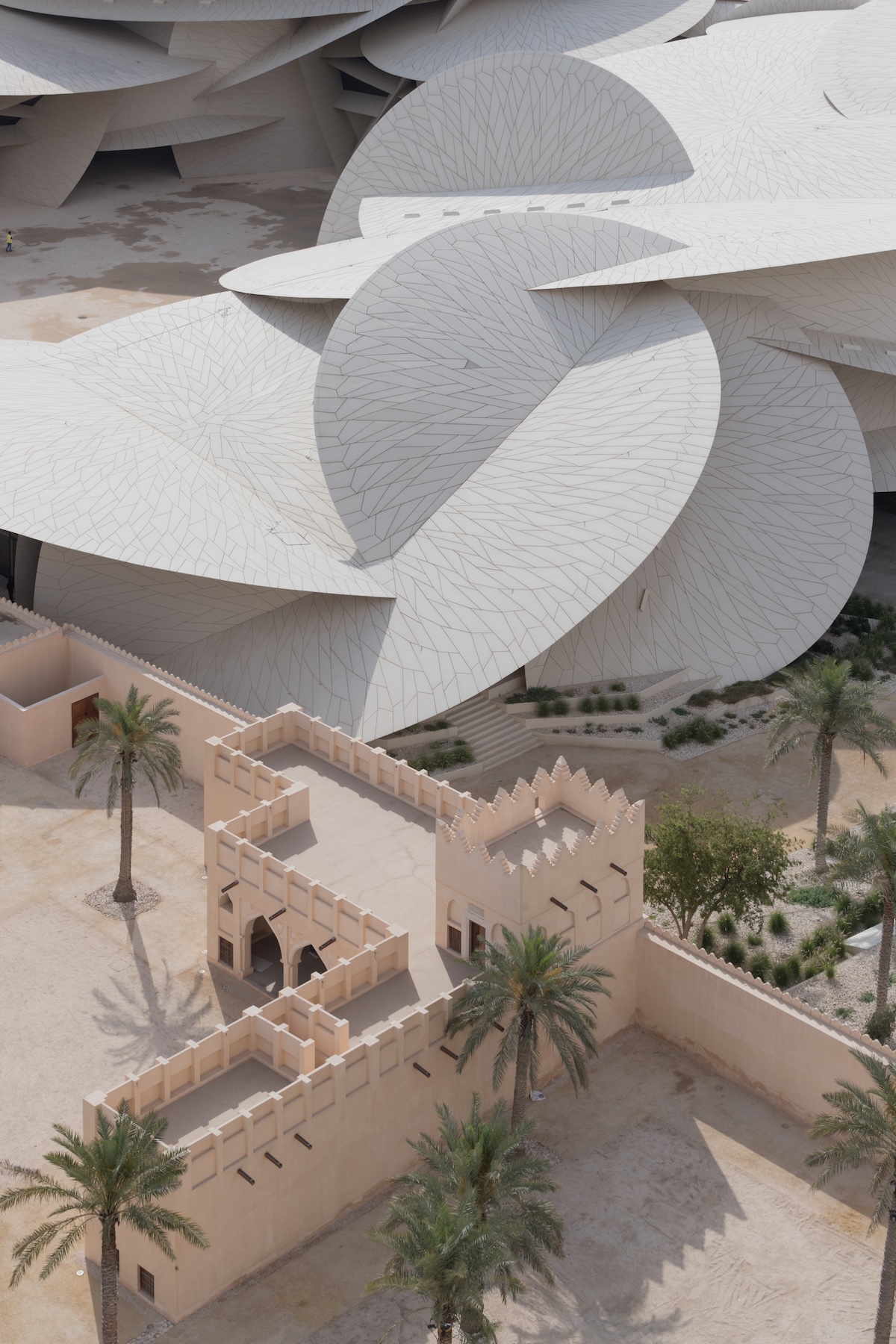 Aerial view of a modern building with overlapping white, petal-like structures positioned next to a traditional sand-colored fort with palm trees in a desert setting.