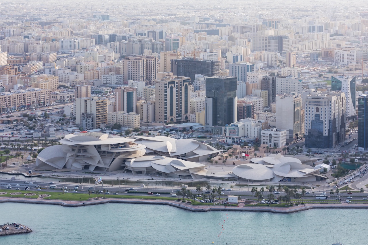 Aerial view of a modern city with a unique, futuristic building featuring overlapping, disc-like structures by a waterfront, surrounded by high-rise buildings and busy roads