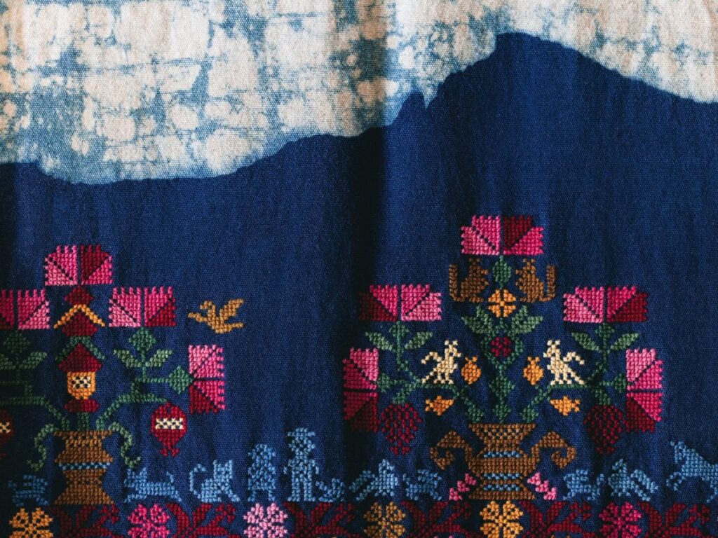 Colorful cross-stitch embroidery with floral and geometric patterns on deep blue fabric, featuring a light blue and white cloud-like design at the top, inspired by the artistic vision of Chef Noof Al Mari.
