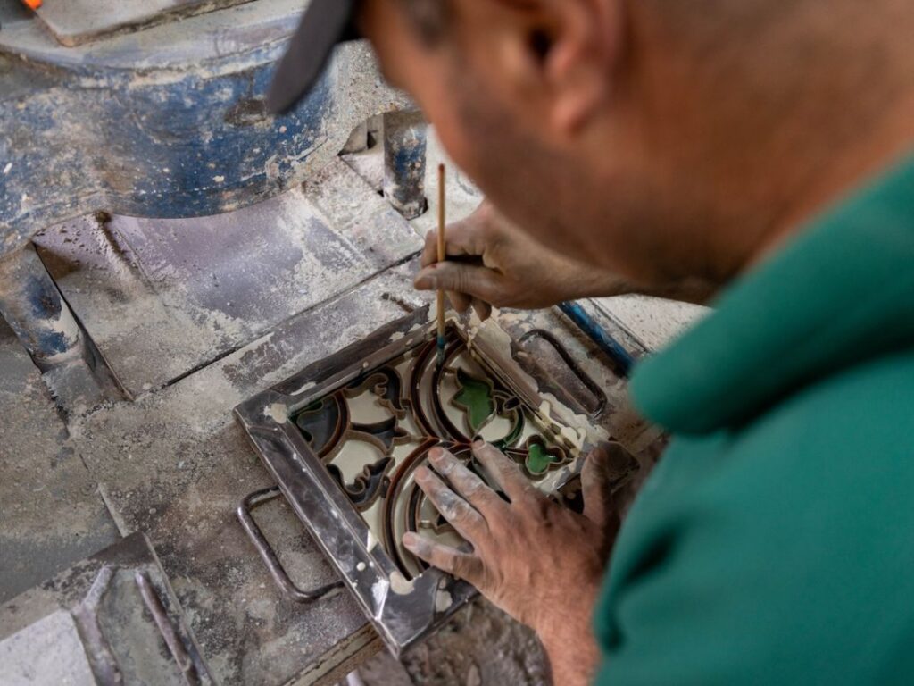 A person uses a small tool to carefully paint designs onto a square ceramic tile, which features intricate patterns in green, brown, and black reminiscent of Frida Escobedo’s distinctive style. Their hands and the surrounding workspace are covered in dust.