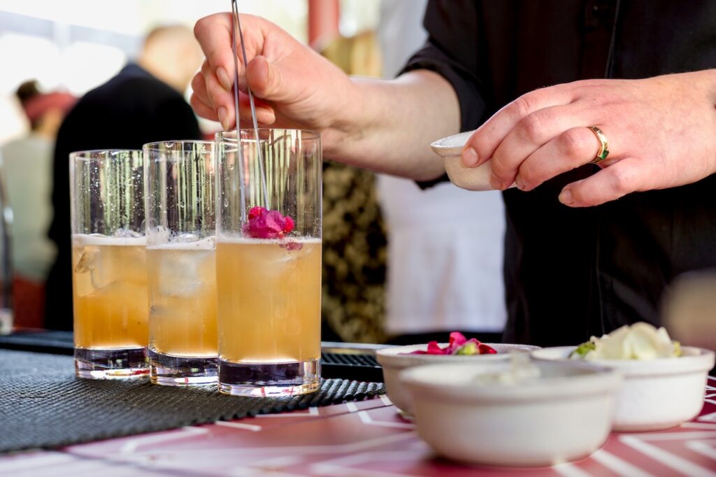 A person garnishes tall glasses of iced cocktails with pink flowers. Nearby bowls hold additional garnishes on a counter.