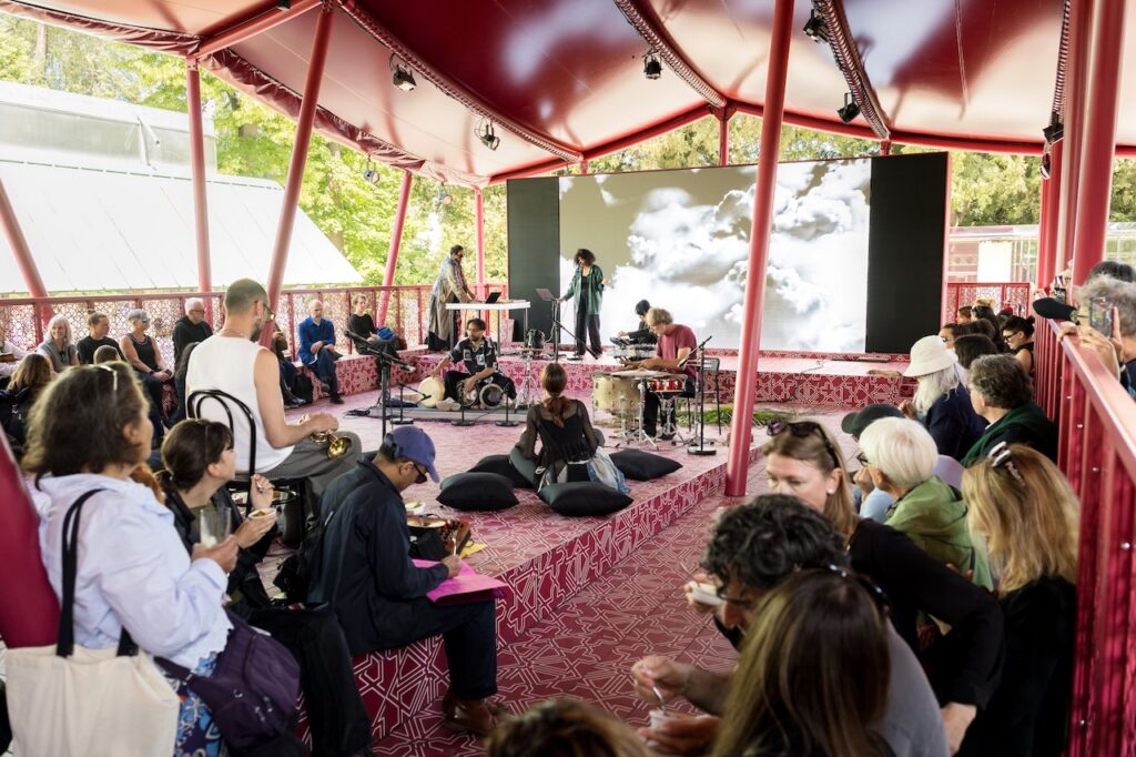 People gather under a red open-air pavilion in Msheireb, some seated on cushions, watching musicians perform on a stage with a cloudy sky backdrop. Daylight filters through trees outside.