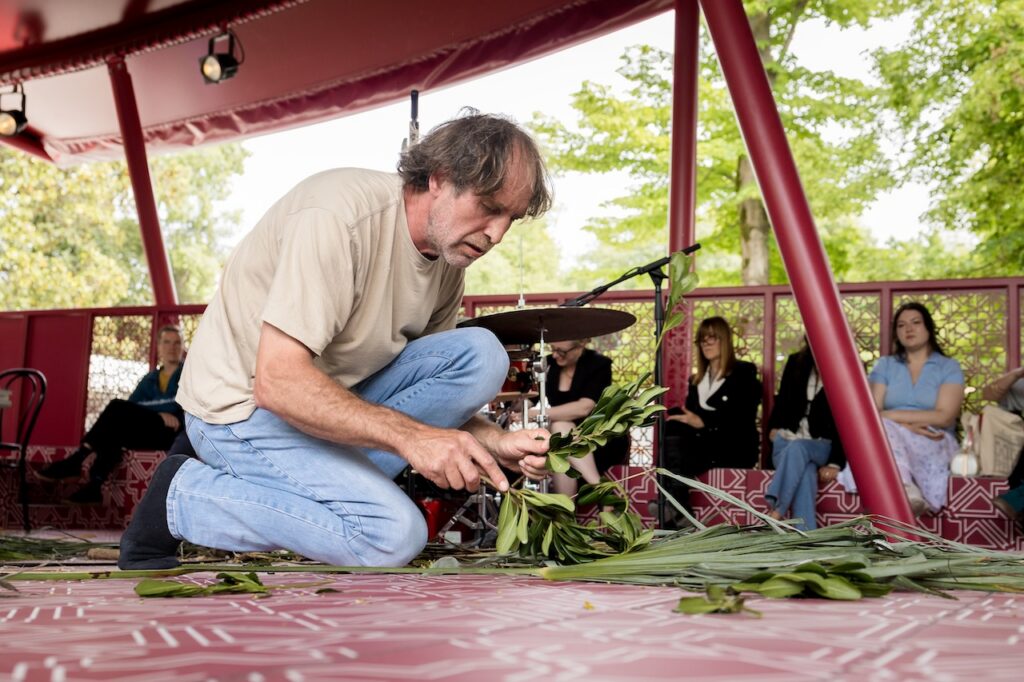 A man kneels on a patterned red floor, handling green leaves, with a drum nearby. Several people sit and watch him in an outdoor pavilion surrounded by trees.