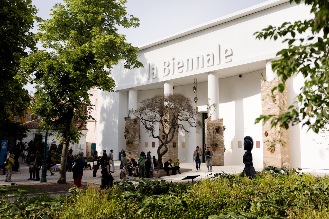 People gather outside a white building with La Biennale written on it, surrounded by trees, plants, and sculptures